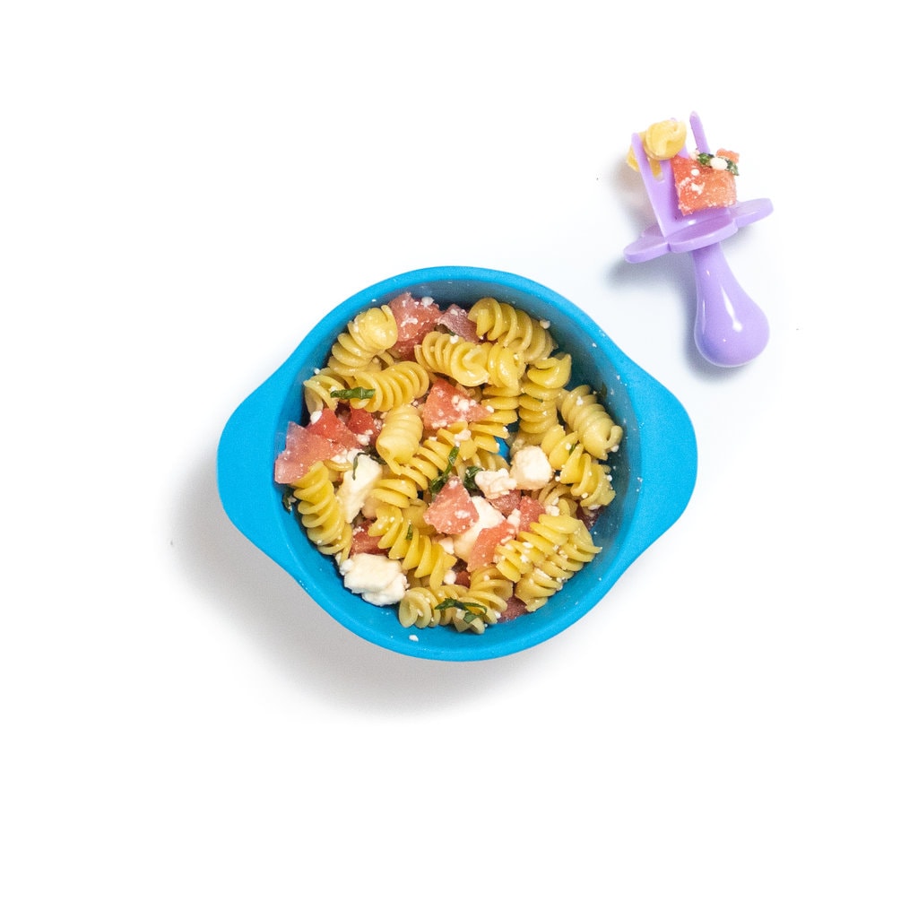 A blue baby bowl filled with a mix of pasta, watermelon, feta and basil against a white background with a purple baby fork sitting to the side.