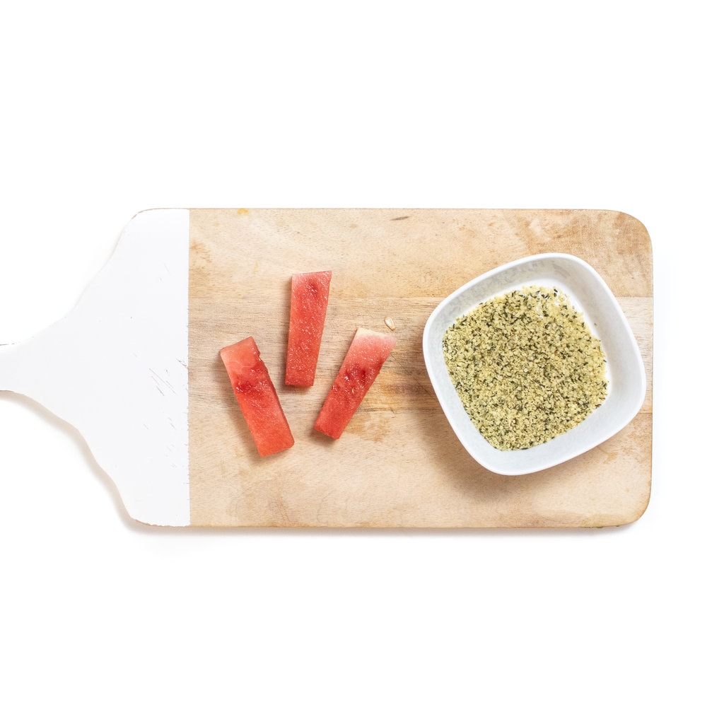 A wooden Korean board with three sticks of watermelon in a small bowl of hemp seeds.