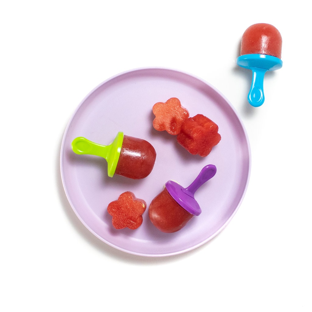 Purple kids plate with colorful popsicle sticks with watermelon popsicles and chunks of watermelon.