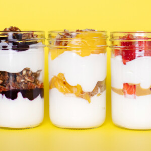 Three glass jars full of fruit filled yogurt parfait's for kids with lots of fruit and healthy toppings against a yellow background.