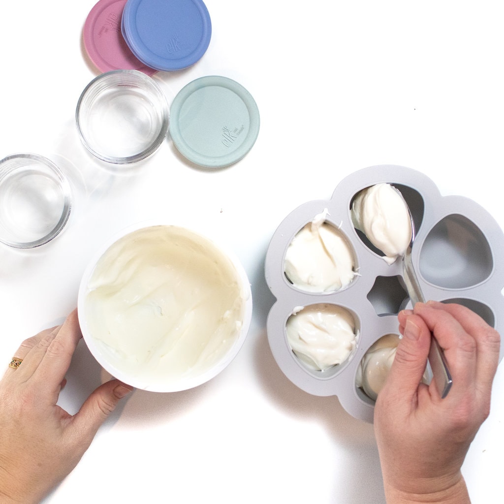 Spread of two hands spooning white yogurt into a ice cube tray with glass containers in lids nearby against a white background.