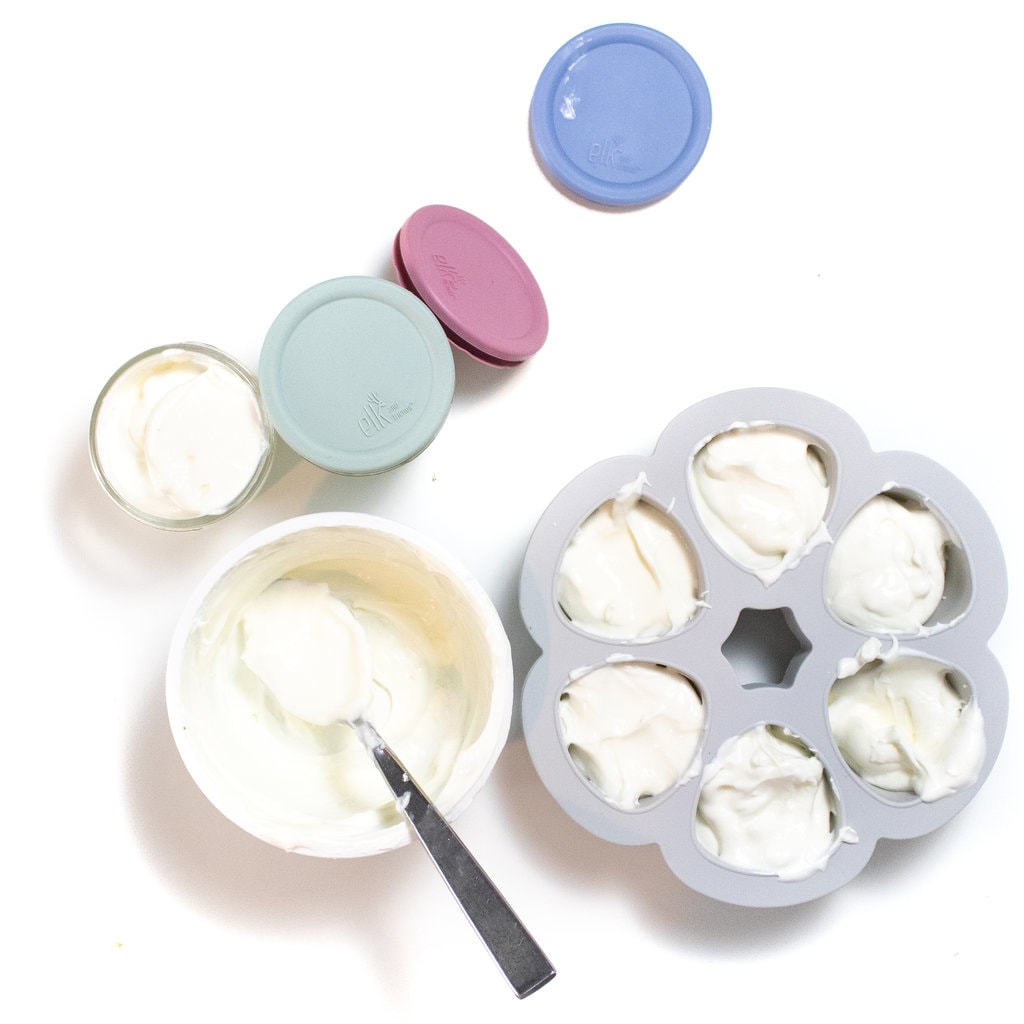 An open container of yogurt that has been spooned into an ice cube tray and clear glass jars with colorful lids.