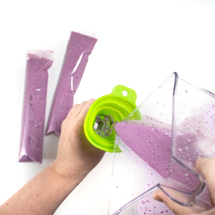 Two hands - one pouring blueberry yogurt from a blender into a clear tube.