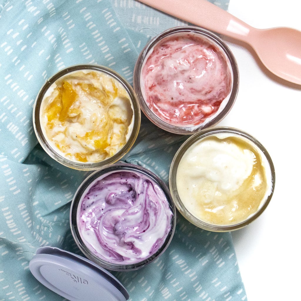 Four glass jars full of yogurt with fruit swirled on top with a blue napkin a pink spoon and a blue lid against a white background.