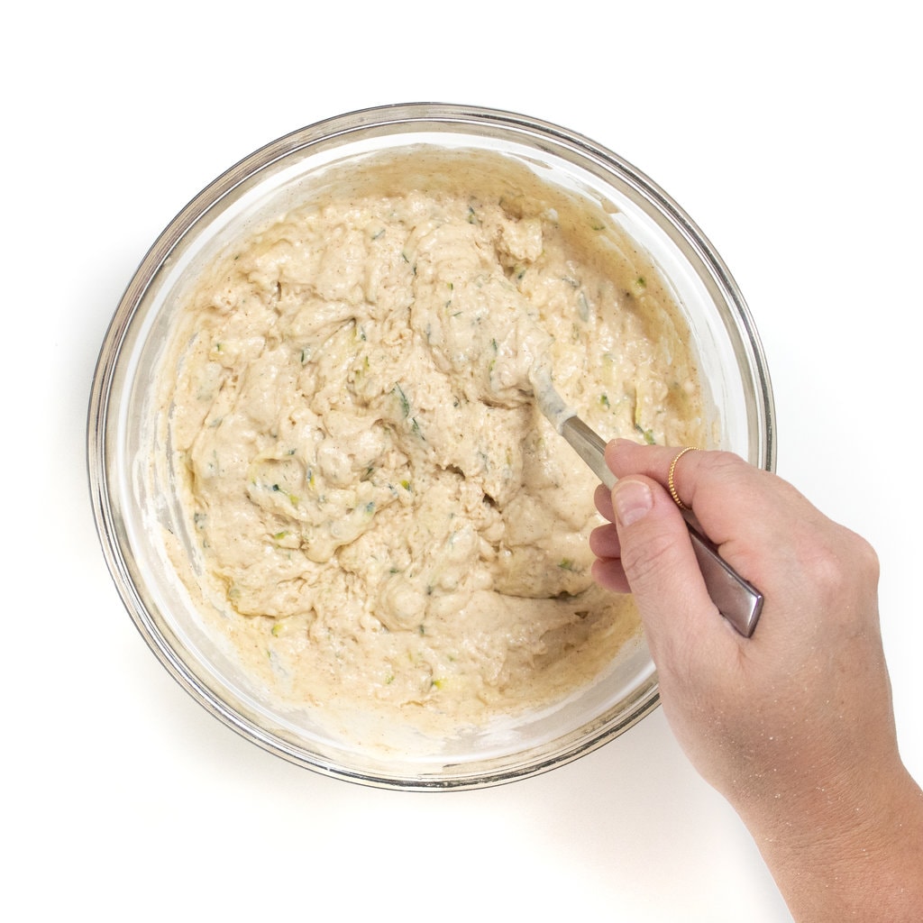 Against a white background with the zucchini pancake batter mixed together with a hand stringing it.