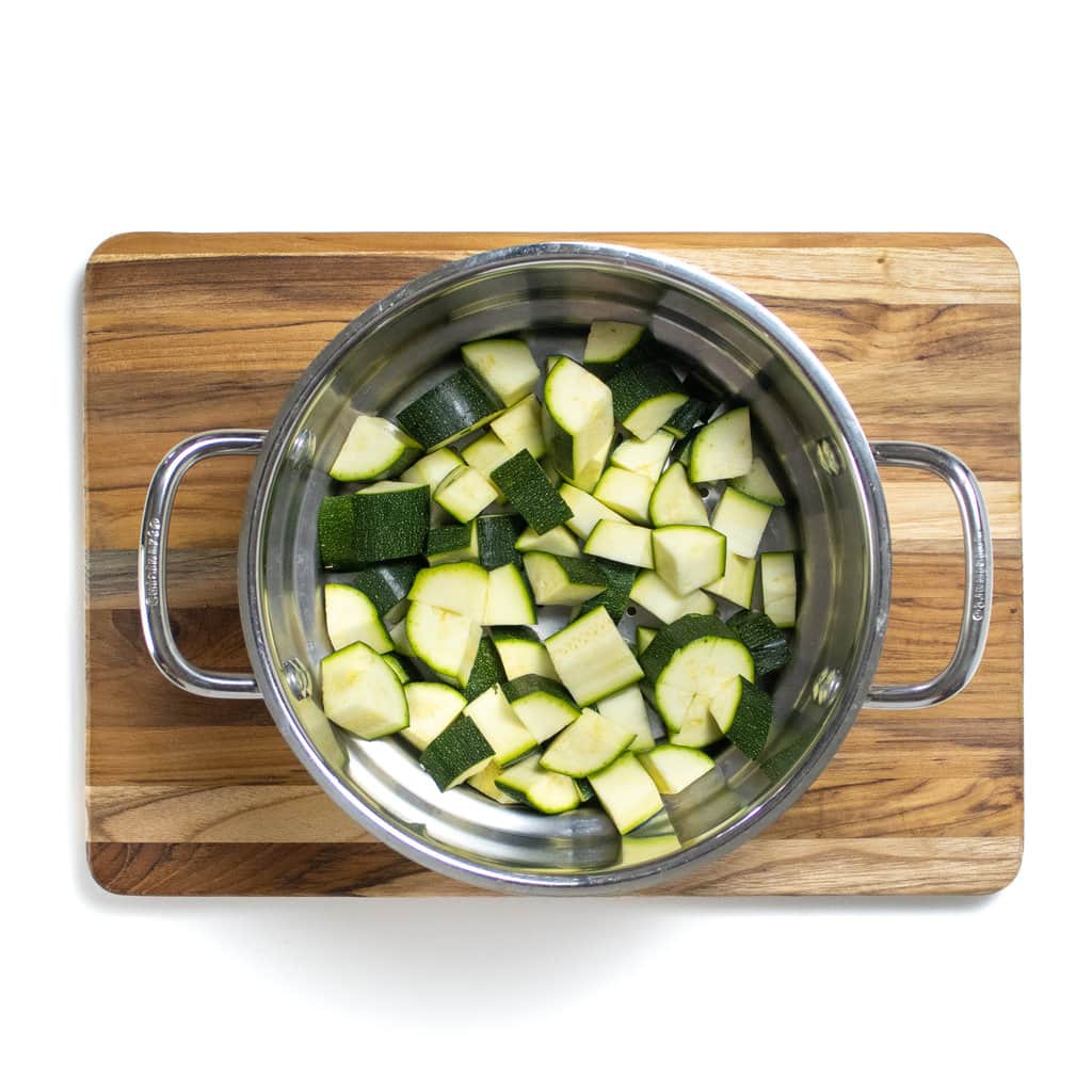 Cut zucchini in a steamer basket sitting on a cutting board.