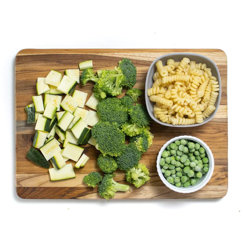 A wooden cutting board with chopped zucchini, broccoli, pasta and peas for a baby food.