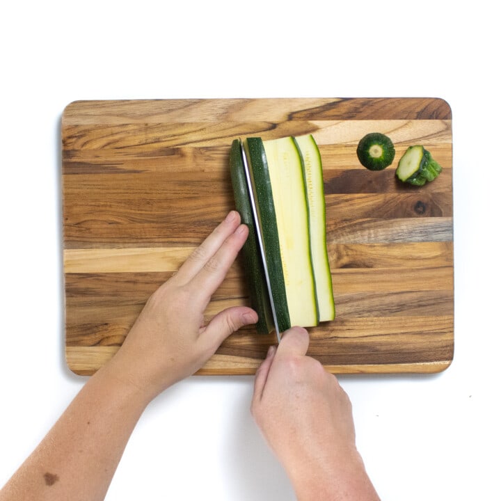 A wooden cutting board with two hands cutting a zucchini into planks.