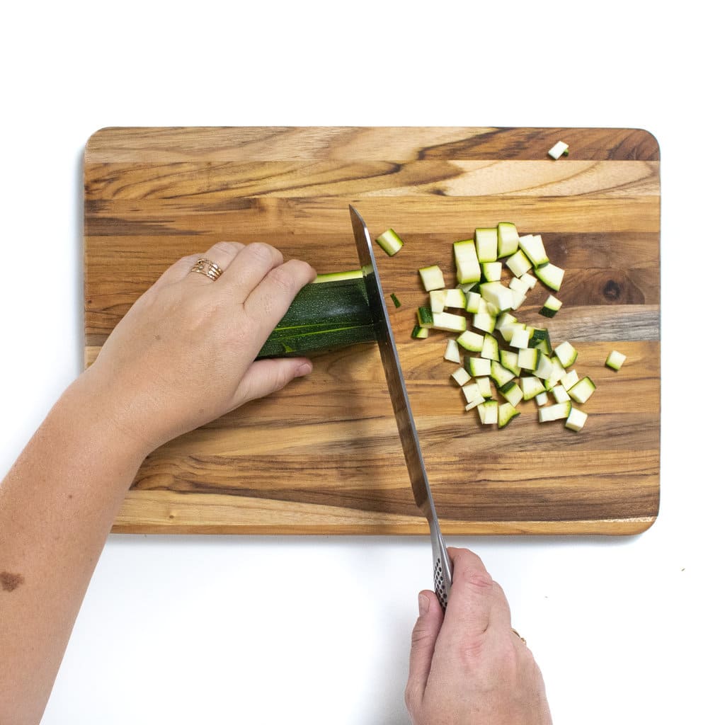 Two hands cutting a zucchini into small pieces on a wooden cutting board against a white background.