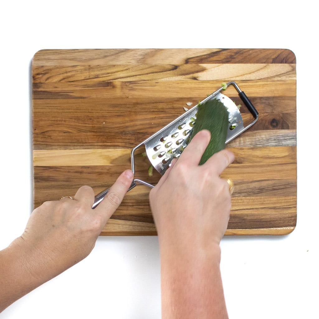 Two hands grading a zucchini over a cutting board.