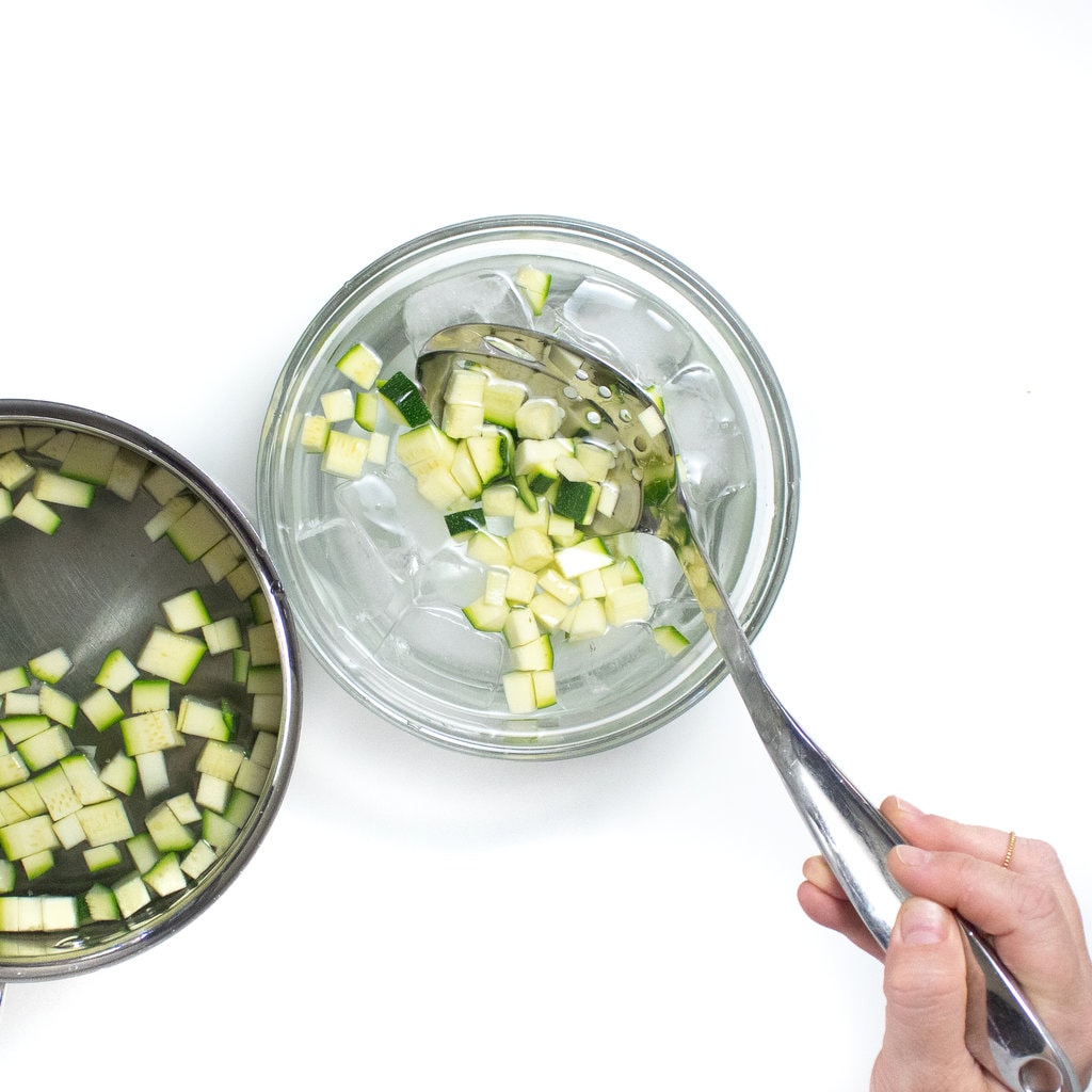 Hand holding a silver spoon transferring cooked zucchini to an ice bath.