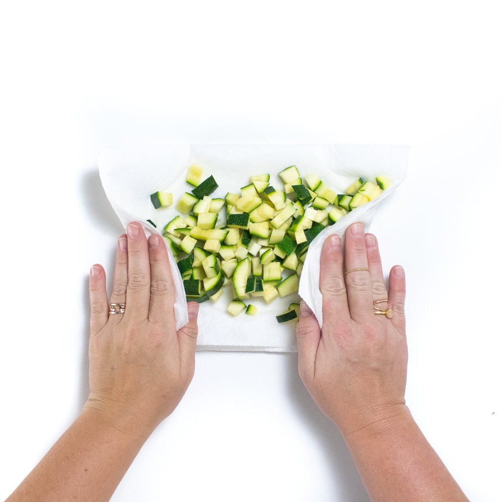 Two hands drying off diced zucchini with a paper towel.