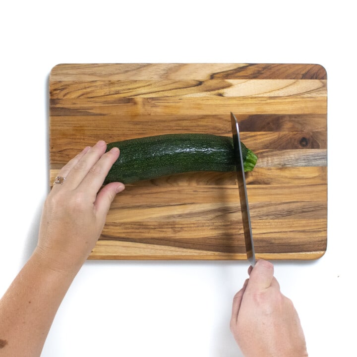 A wooden cutting board with two hands cutting a zucchini into slices.