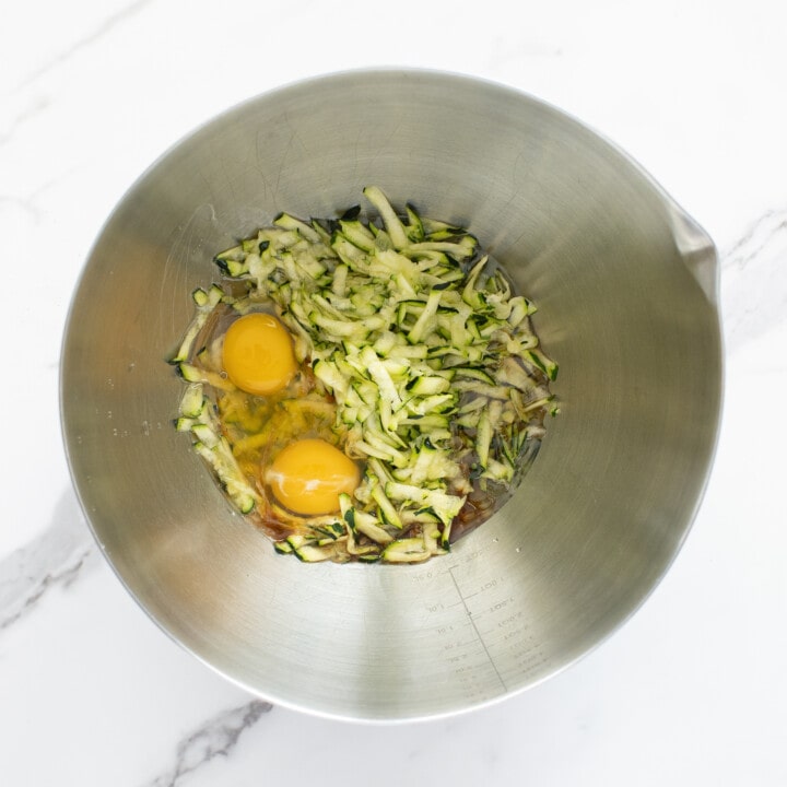 Silver mixing bowl on a marble counter filled with zucchini and other wet ingredients.