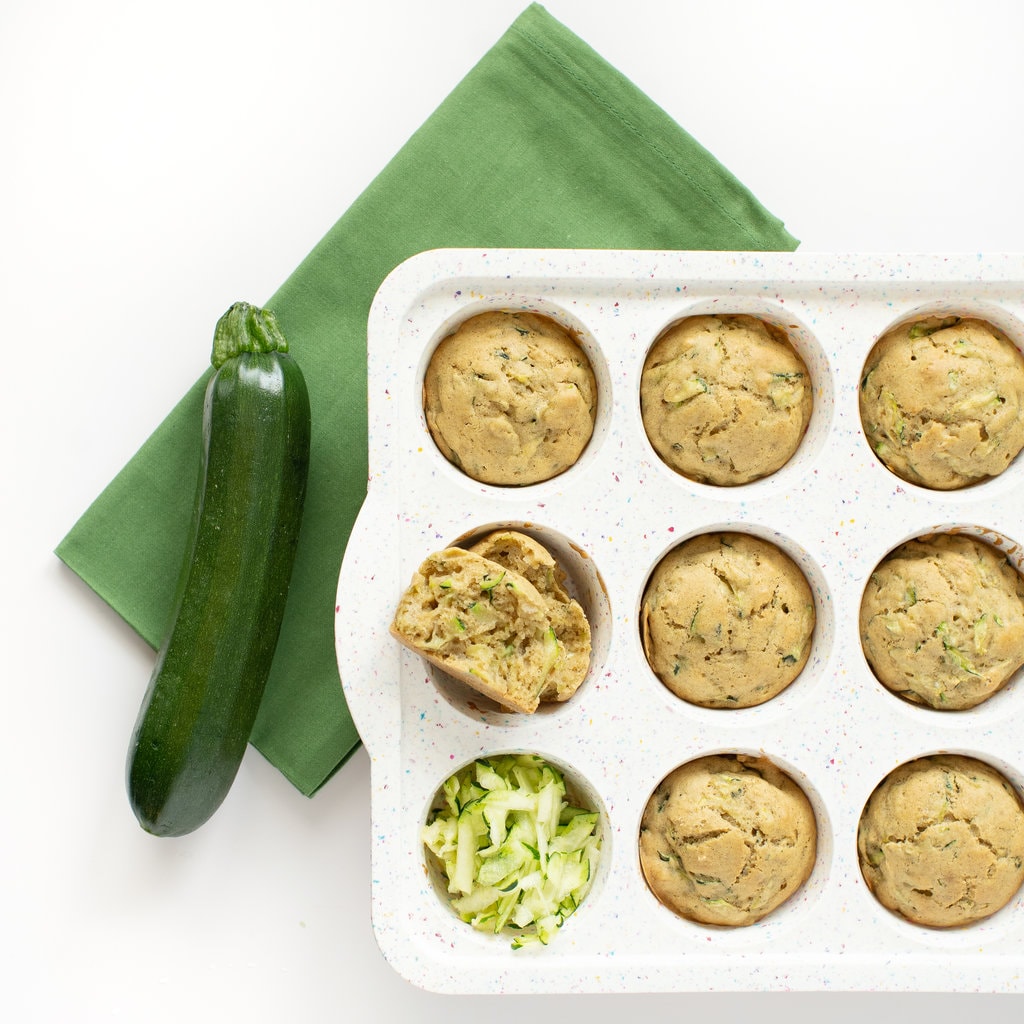 White muffin tin with cooked zucchini muffins on a green napkin with a zucchini sitting next to it and shredded zucchini in one of the muffin rings.