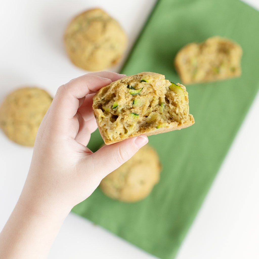 Small hand holding a muffin over a table with muffins and a green napkin.